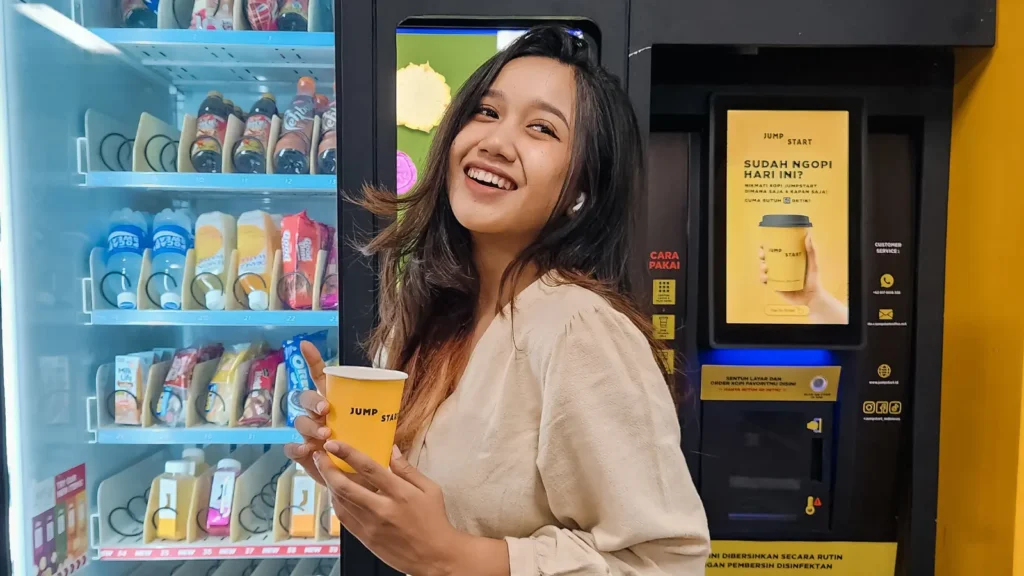 Seorang wanita memegang kopi dari mesin JumpStart sambil tersenyum di depan vending machine pintar.